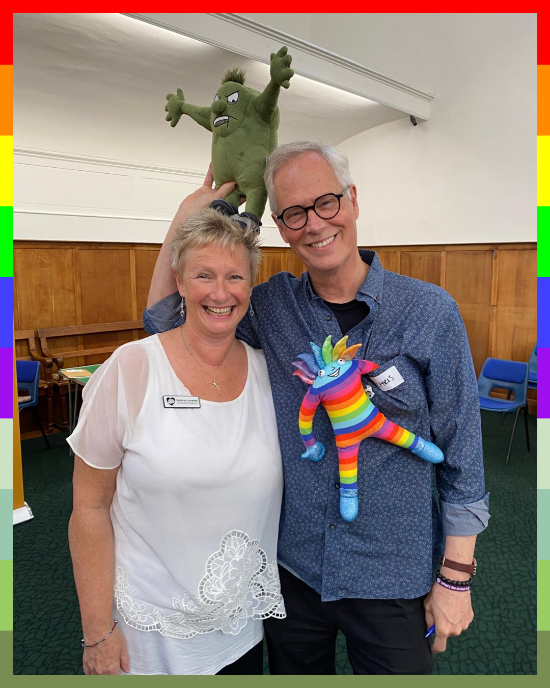 Kathryn Lovewell and Chris Germer are holding colorful plush toys, Booster and Crusher, in a room with wooden paneling.