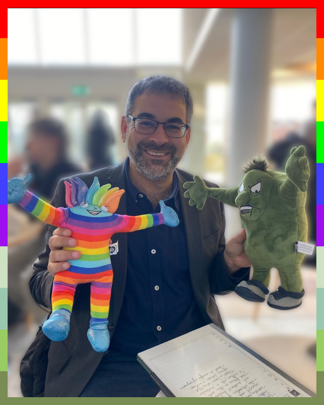 Professor Robin Banarjee holding a rainbow-colored plush toy, Booster and a green plush toy Crusher, in an indoor setting